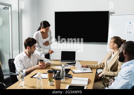 donna d'affari afro-americana incinta che punta al laptop vicino ai colleghi Foto Stock