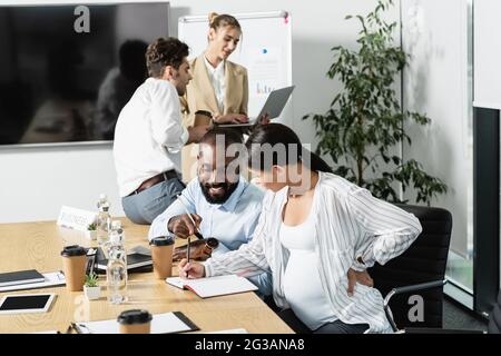 sorridente afroamericano che punta al notebook vicino donna incinta d'affari e colleghi parlanti Foto Stock