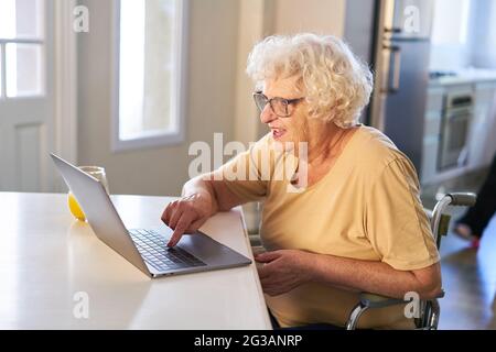 La donna anziana si diverte sul computer portatile mentre chiacchiera o scrivi e-mail nel suo appartamento Foto Stock