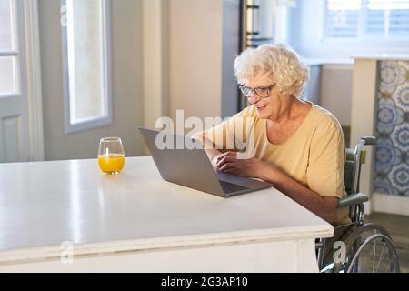 Donna anziana felice in sedia a rotelle Video Chat con la famiglia su computer portatile a casa Foto Stock