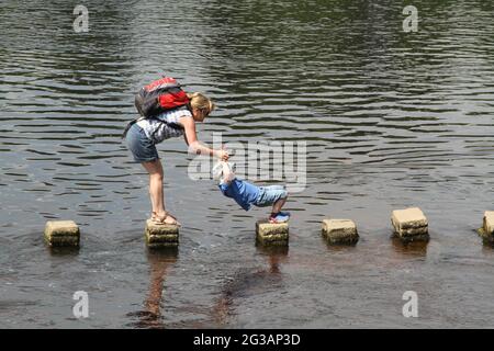 Abbazia di Bolten, Skipton, Yorkshire, Inghilterra. Una mamma giovane aiuta suo figlio a incrociare l'uso di pietre che corrompono il fiume Wharfe Foto Stock