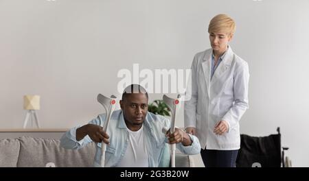 Felice giovane infermiera fornisce assistenza, assistenza medica, aiuto e supporto. Donna adulta seria guarda l'uomo afroamericano maturo con stampelle alzarsi Foto Stock