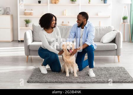 Felice famiglia d'amore. Ritratto di belle spose afroamericane che pattugano il cane seduto sul pavimento moquette in appartamento moderno, guardando l'un l'altro. SM Foto Stock