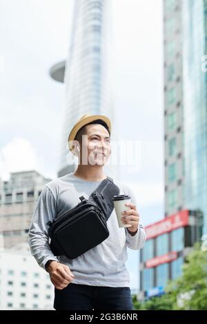 Positivo bell'uomo asiatico in cappello da sole e auricolari wireless che tengono una tazza di caffè e che cammina nella grande città Foto Stock