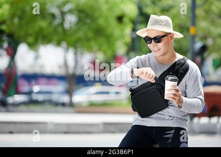 Positivo giovane turista vietnamita in cappello e occhiali da sole seduti contro la strada della città e controllare il tempo mentre si beve caffè in estate Foto Stock