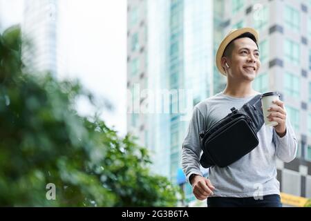 Allegro giovane turista vietnamita con cintura nera a piedi con caffè da asporto in grande città e guardando intorno Foto Stock