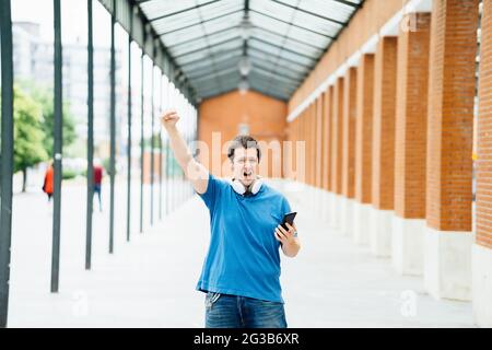 Giovane adulto che controlla smartphone esprimendo successo clenching suo pugno alzando il braccio felice in strada gridando. Concetto ricevere buone notizie Foto Stock