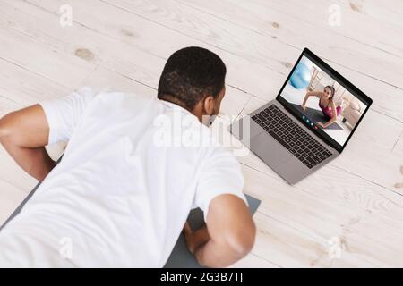 Black Young Man fare formazione online con il laptop a casa, irriconoscibile guy afroamericano fare esercizio Plank mentre si dispone di videochiamata con FEMA Foto Stock