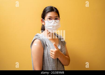 Vaccinato ragazza teen asiatica in maschera di faccia che mostra bendaggio intonaco sul suo braccio dopo aver ottenuto l'iniezione di vaccino covid-19 su sfondo giallo studio. Coron Foto Stock