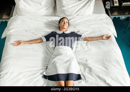 Felice giovane cameriera in uniforme sdraiata su un grande letto matrimoniale Foto Stock