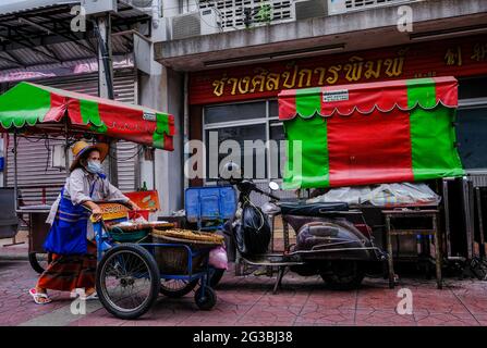 Un'anziana venditrice di strada femminile, che vende noci, spinge il suo carrello davanti ad alcune colorate tettoie a Chinatown, Bangkok, Thailandia. Foto Stock