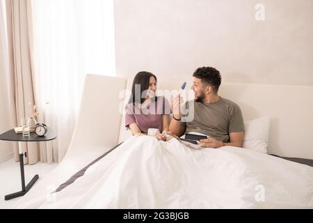 Donna felice che guarda il suo marito surpwised con il test di gravidanza mentre si sdraia a letto Foto Stock