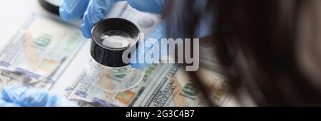 Mani in guanti di gomma che tengono la lente di ingrandimento sopra le fatture del dollaro primo piano Foto Stock