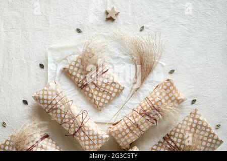 Regali fatti a mano avvolti in carta da artigianato beige con erba secca selvatica legata con filo di cotone. Piatto geometrico steso su tessuto bianco con decorazione naturale Foto Stock