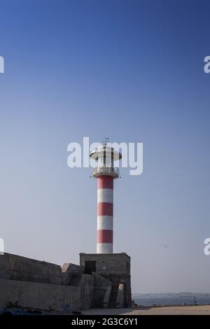 Una vista del porto con un grande vecchio faro rosso bianco, molo contro il cielo blu e il mare Foto Stock