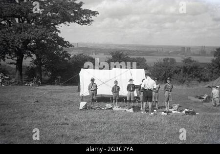 1951, storico, all'esterno in un campo di erba, da una tenda, scouts giovani in uniforme cucciolo, in piedi in una linea che ha un kit di ispezione da parte del maestro scout, Inghilterra, Regno Unito. Foto Stock