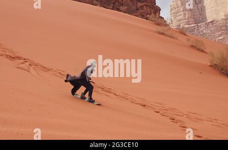 Giovane uomo sabbia duna surf con bisht - cappotto beduino tradizionale. Il Sandsurfing è una delle attrazioni del deserto di Wadi Rum Foto Stock