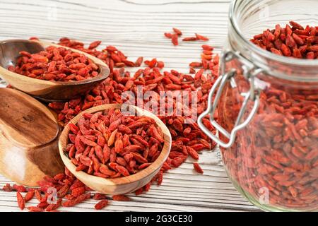 Goji secco aka. Semi di mirtillo all'interno di una bottiglia di vetro, alcuni in ciotole di legno e versato sulla scrivania delle tavole bianche Foto Stock