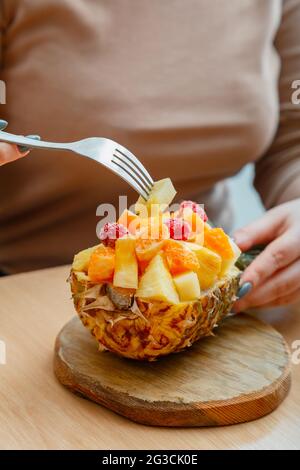 Donna che mangia gustosa insalata di frutta tropicale in mezza ciotola di ananas. Frutti di bosco lamponi ananas mandarino arance in insalata di frutta da dessert in caffetteria. Cibo Foto Stock