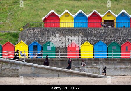 Una famiglia discende sulla spiaggia per godersi le delizie del mare britannico davanti a una fila di colorate capanne sulla spiaggia di Whitby nel Nord Yorkshi Foto Stock