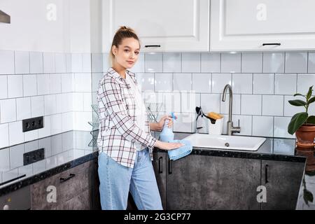 Donna pulizia cucina con spray detergente e panno piatto, concetto per l'igiene. Donna bionda in abiti casual pulizia moderna cucina, guardare venuto Foto Stock