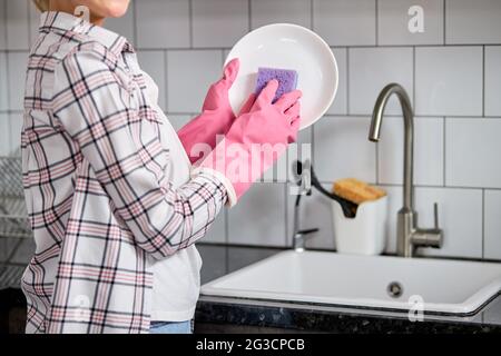 Piatti lavanti femmina corti con spugna viola sopra lavello della cucina , casalinga irriconoscibile a casa facendo i chores della famiglia, indossando gomma Foto Stock
