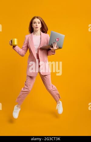 Preoccupata bella donna allegra leader di successo saltando in aria trasportando il laptop e la tazza di caffè in esecuzione veloce fretta-up riunione isolato sopra il colore giallo Foto Stock