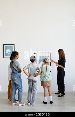 Vista verticale sul retro in un gruppo diverso di bambini, ascoltando la guida del tour femminile mentre visiti la galleria d'arte moderna Foto Stock