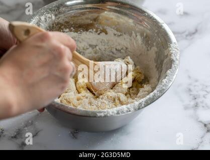 donna mano che tiene un cucchiaio di legno mescolando gli ingredienti in una ciotola rotonda di metallo su una superficie di marmo bianco Foto Stock