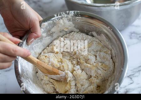 donna che tiene un cucchiaio di legno mescolando gli ingredienti in un recipiente rotondo di metallo angolato su una superficie di marmo bianco Foto Stock