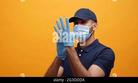 Uomo di consegna che indossa i guanti. Indossare una maschera e un cappuccio blu prendendo precauzioni. Concetto di Covid 19 e Pandemic. Foto di studio con sfondo giallo. Foto Stock