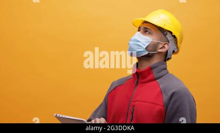 Un giovane ingegnere che indossa un casco di sicurezza giallo e una maschera che tiene in mano una tavoletta. Studio con sfondo giallo. Progettazione e costruzione. Foto Stock
