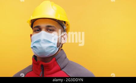 Ritratto di un giovane ingegnere indiano che indossa un casco giallo di sicurezza e una maschera in piedi nello studio con sfondo giallo. Uomo isolato. Espressione facciale positiva. Primo piano. Foto Stock