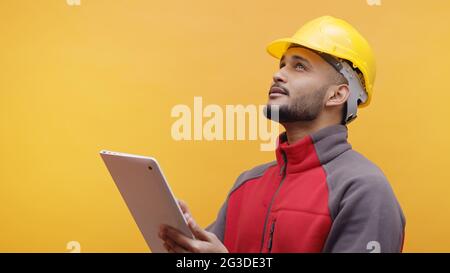 Un giovane ingegnere che indossa un casco di sicurezza giallo che tiene una tavoletta in mano. Studio con sfondo giallo. Progettazione e costruzione. Foto Stock