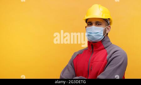 Ritratto di un giovane ingegnere indiano che indossa un casco giallo di sicurezza e una maschera in piedi con le braccia incrociate nello studio con sfondo giallo. Uomo isolato. Primo piano. Foto Stock