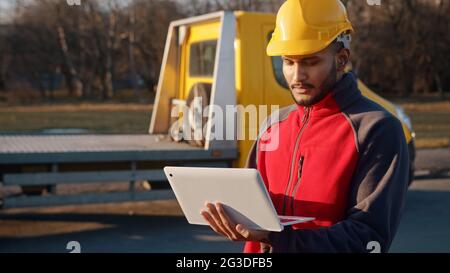 Giovane ingegnere che indossa una divisa e un casco di sicurezza in piedi con un camion giallo dietro di lui. Tenendo un computer portatile in mano. Concetto di ingegneria e costruzione Foto Stock