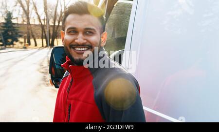 Il ragazzo di consegna in piedi sulla strada sorridente. Dietro di lui si può vedere un furgone bianco. Ragazzo di consegna che indossa uniforme rossa con espressioni facciali positive. Alberi sullo sfondo. Luce del sole che riflette il suo volto. Foto Stock