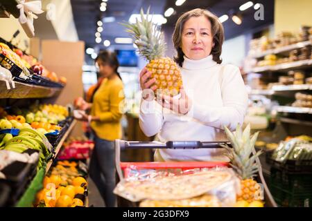 Donna anziana soddisfatta che raccoglie ananas maturo al supermercato Foto Stock