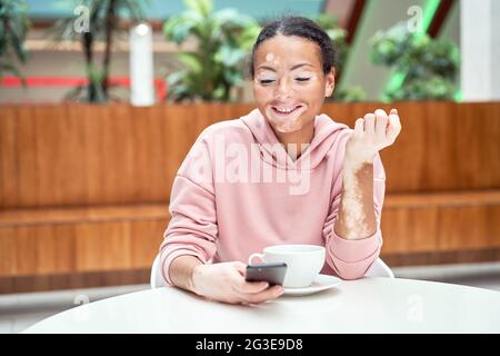 Donna nera afroamericana con pigmentazione vitiligina problema della pelle interna tavolo con felpa con cappuccio rosa vestito con smartphone Foto Stock
