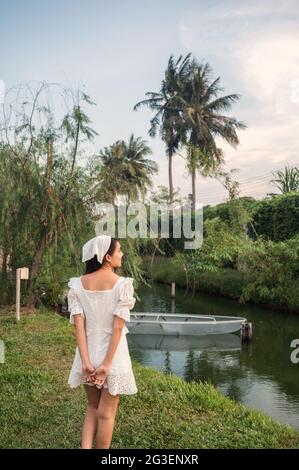 Allegra bella giovane donna asiatica in abito bianco in piedi vicino al canale nel giardino Foto Stock