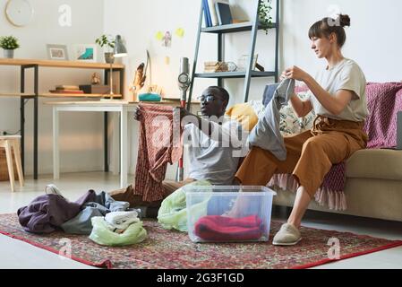 Giovane coppia multietnica seduta sul pavimento e mettendo i vestiti in ordine nelle scatole a casa Foto Stock