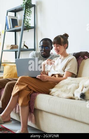 Giovane coppia multietnica seduta sul divano guardando qualcosa sul computer portatile e sorridendo che trascorrono il tempo a casa Foto Stock
