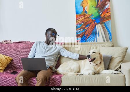 Giovane africano che strinse il suo cane mentre si siede sul divano e lavora online sul computer portatile nella stanza Foto Stock