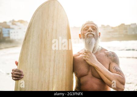 Surfista senior tatuato che tiene una tavola da surf vintage sulla spiaggia Al tramonto - fuoco sul viso Foto Stock