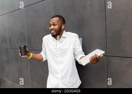 Giovane uomo afro-americano uscito in abiti casual in piedi all'aperto sorridendo, bevendo una tazza di caffè take away e tenendo un tablet digitale in città Foto Stock