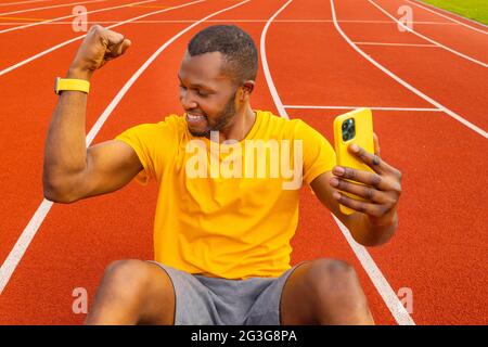 Bello sportivo Afro americano millennial maschio in casual giallo t-shirt sorridente felicemente e alzando pugno clenched, tendere muscoli, sentire forte e. Foto Stock