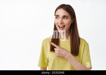 Primo piano ritratto di sorridente sorprende 20s ragazza, gastping affascinato con qualcosa di fresco, puntando il dito a sinistra al logo, mostrando una pubblicità impressionante Foto Stock