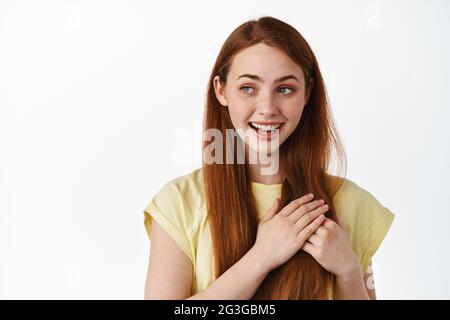 Primo piano ritratto di donna rossa che tiene le mani sul cuore, guardando sinistra sognante, pensando a qualcosa di romantico o amore, in piedi su sfondo bianco Foto Stock