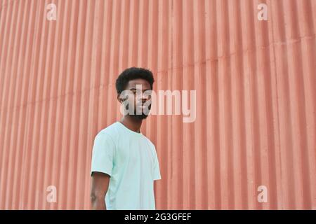 Ritratto di giovane uomo alato africano che guarda la macchina fotografica mentre si trova all'aperto contro il muro rosso Foto Stock