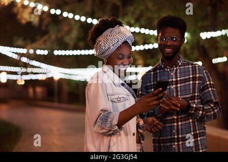 Giovane coppia africana che usa il telefono cellulare insieme mentre si trova nel parco in serata Foto Stock